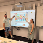 Ein Mann und eine Frau stehen vor einem Whiteboard. Sie zeigen auf eine Grafik mit Comicfiguren und dem Schriftzug "Trau Dich".