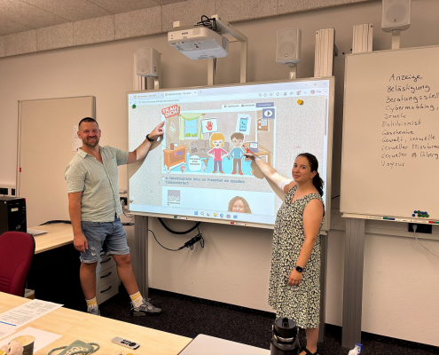 Ein Mann und eine Frau stehen vor einem Whiteboard. Sie zeigen auf eine Grafik mit Comicfiguren und dem Schriftzug "Trau Dich".