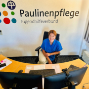 Eine Frau sitzt hinter ihrem Schreibtisch. Hinter hier an der Wand das bunte Paulinenpflege-Logo und der Schriftzug "Jugendhilfeverbund"
