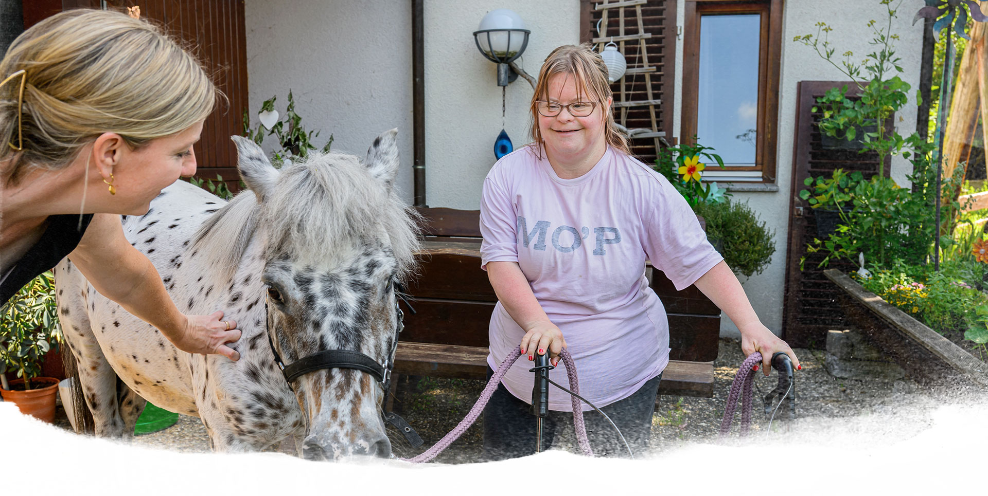 Junge Frau mit Down-Syndrom steht mit ihrem Rollator neben einem gescheckten Pony und hält dessen Führstrick, während eine Betreuerin das Pony streichelt.