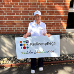 Michiko Pubanz mit Paulinenpflege-Cap und Paulinenpflege-Poloshirt steht vor einer Backsteinwand. Sie hält das Schild mit der Aufschrift "Paulinenpflege - weil das Leben bunt ist" in der Hand.