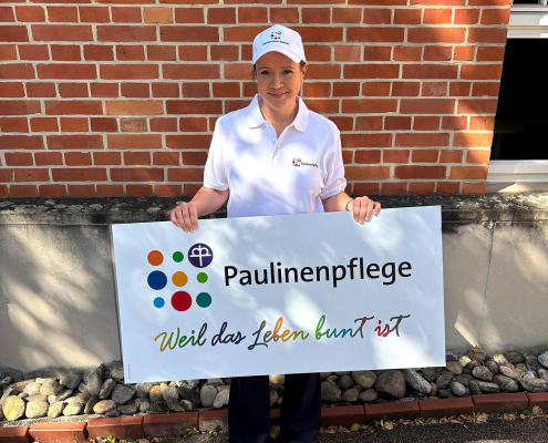 Michiko Pubanz mit Paulinenpflege-Cap und Paulinenpflege-Poloshirt steht vor einer Backsteinwand. Sie hält das Schild mit der Aufschrift "Paulinenpflege - weil das Leben bunt ist" in der Hand.